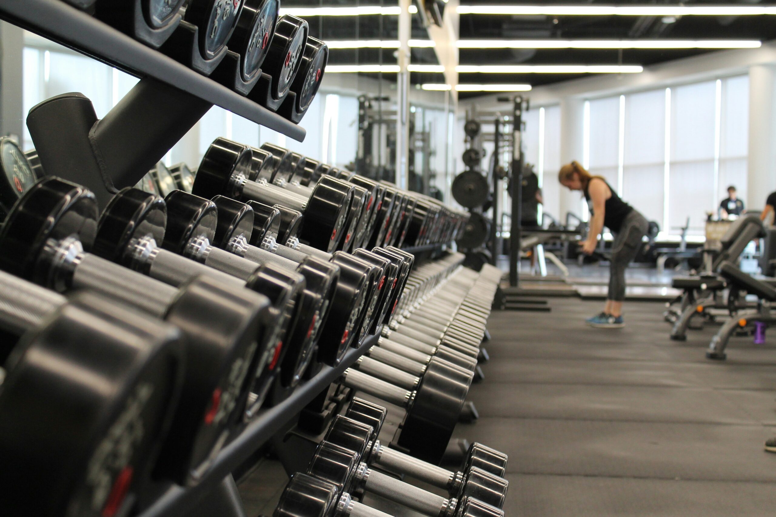 A row of dumbbells in a well-lit gym, with a person in the background lifting weights, symbolizing the role of resistance training in fat loss and muscle development.